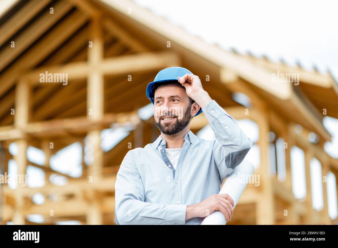 Portrait of an architect or builder in hard hat standing in front of the wooden house structure. Building and designing wooden frame house concept Stock Photo