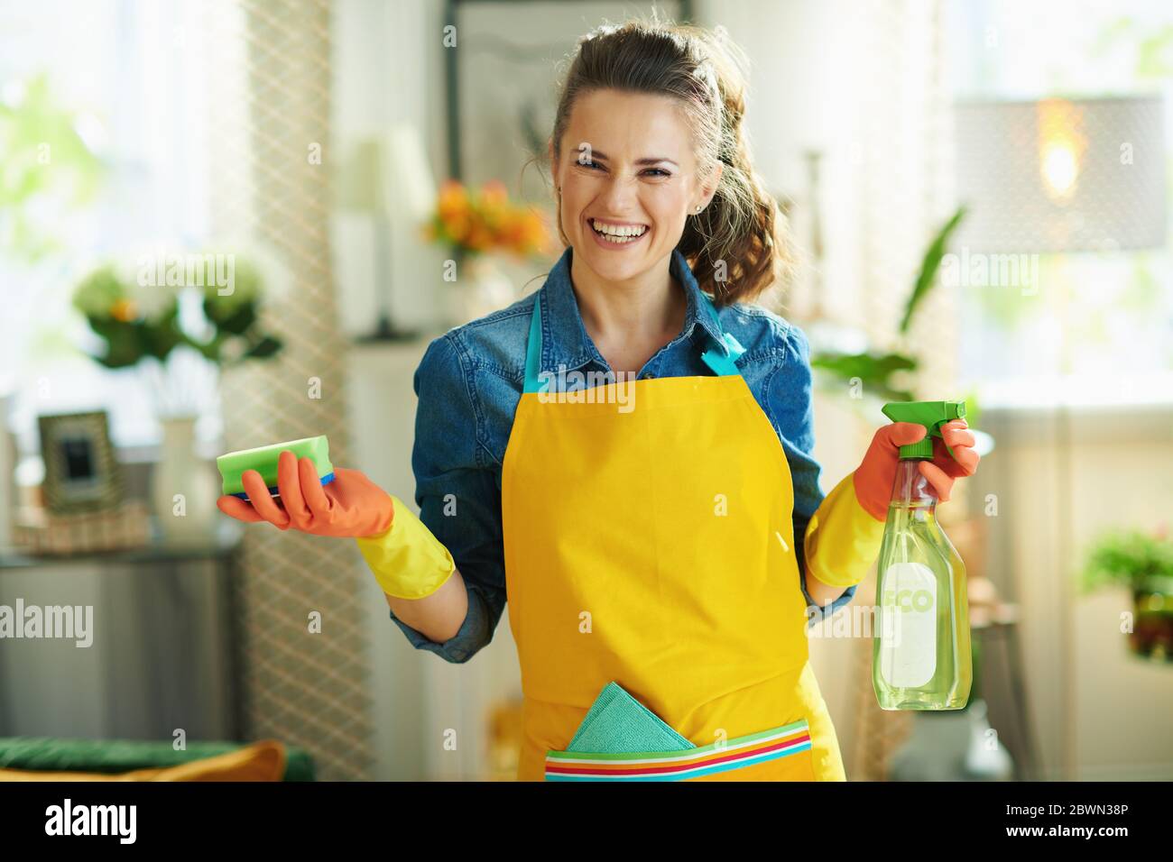 Portrait of happy modern 40 years old woman in orange apron and pink rubber gloves with spray bottle of eco friendly cleaning supplies and green spong Stock Photo