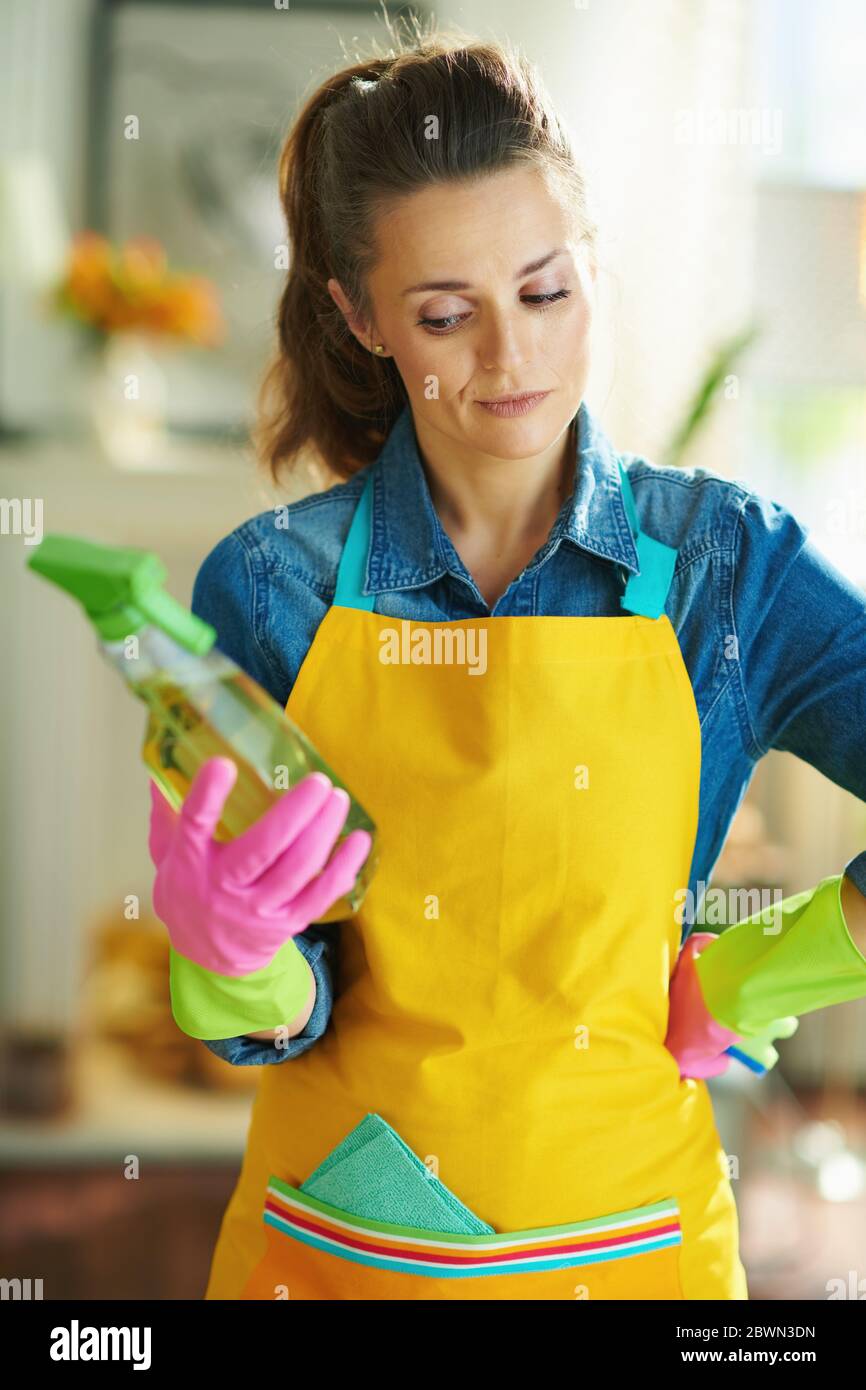 modern housewife in orange apron and pink rubber gloves with spray bottle of cleaning supplies reading label at modern home in sunny day. Stock Photo