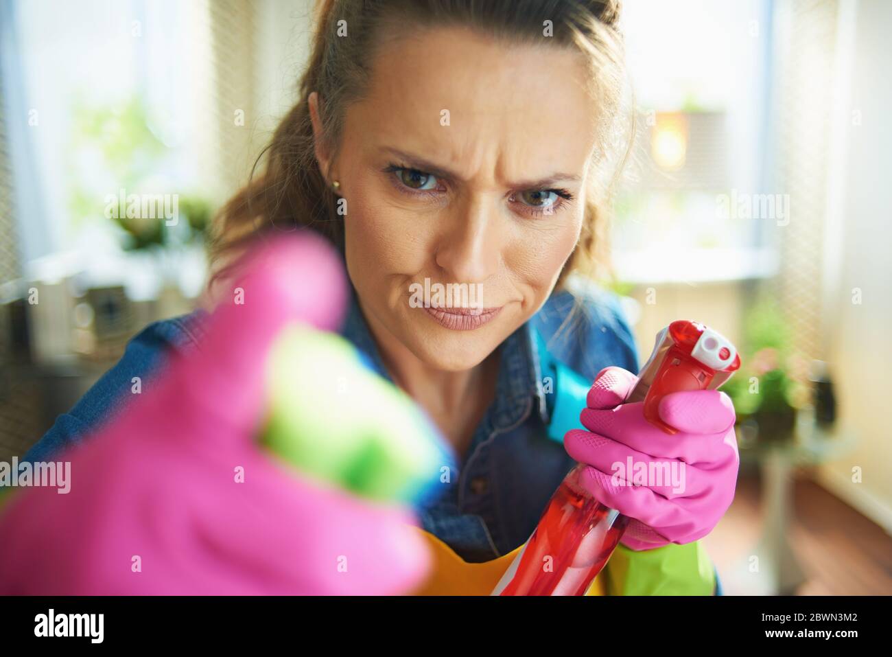 furious modern female in orange apron and pink rubber gloves with spray bottle of cleaning supplies threatening with finger in the modern house in sun Stock Photo