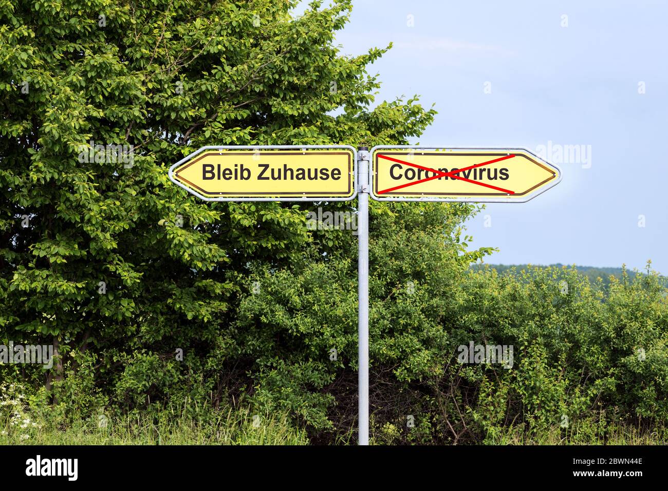 Yellow road signs pointing in opposite directions with German text Bleib Zuhause meaning Stay Home,  and Coronavirus, rural landscape in the backgroun Stock Photo