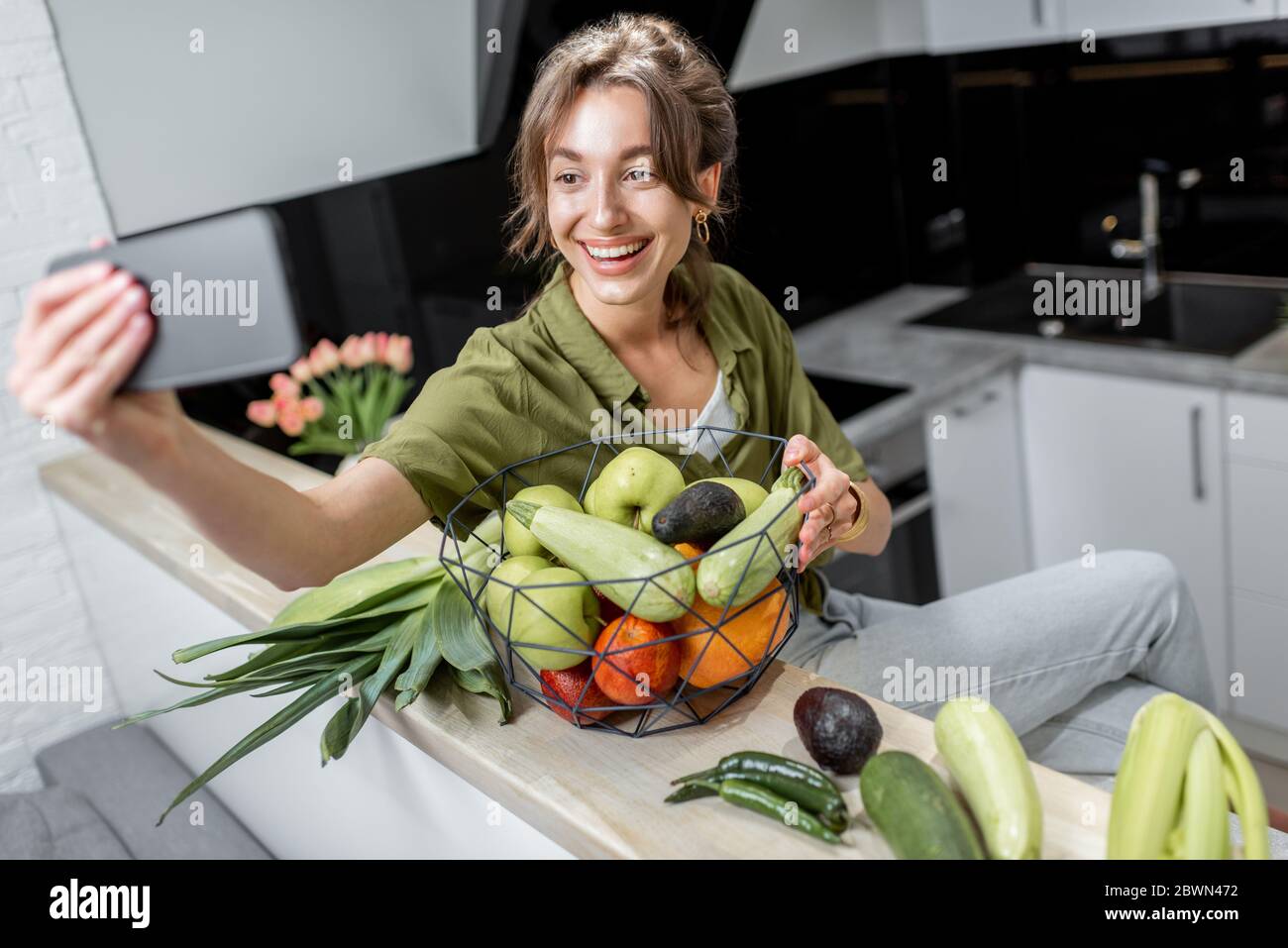 Young woman making selfie photo or vlogging on mobile phone about healthy eating, sitting with raw ingredients on the kitchen at home. Healthy lifestyle and social influence concept Stock Photo