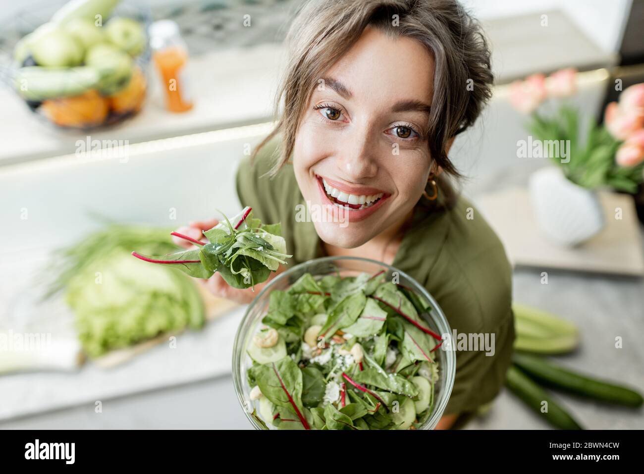 Portrait of a young and cheerful woman eating salad standing on the kitchen with food ingredients on the background, view from above. Vegetarianism, wellbeing and healthy lifestyle concept Stock Photo