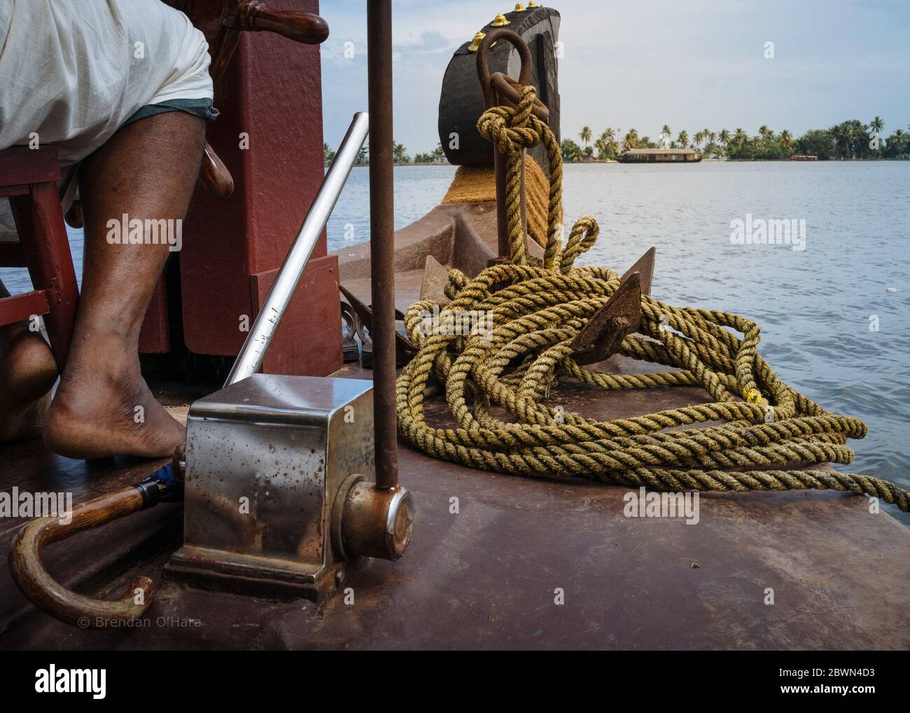 Anchor and umbrella rest at the feet of a kettuvallam skipper as the bow points across the water at the palm tree lined horizon in Kerala's backwaters. Stock Photo