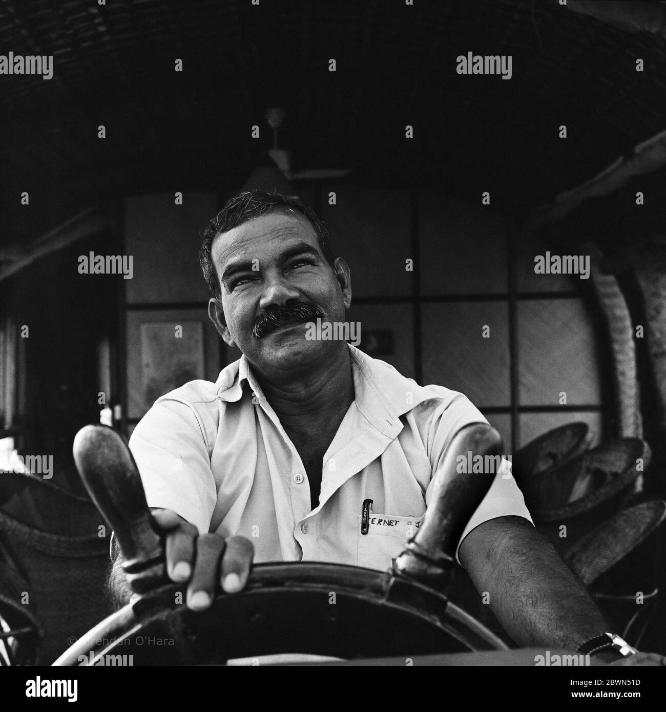 Smiling, mustachioed, doe-eyed boat skipper casualy handels the ship wheel to keep the Kettuvallam on course in Kerala's backwaters. Stock Photo