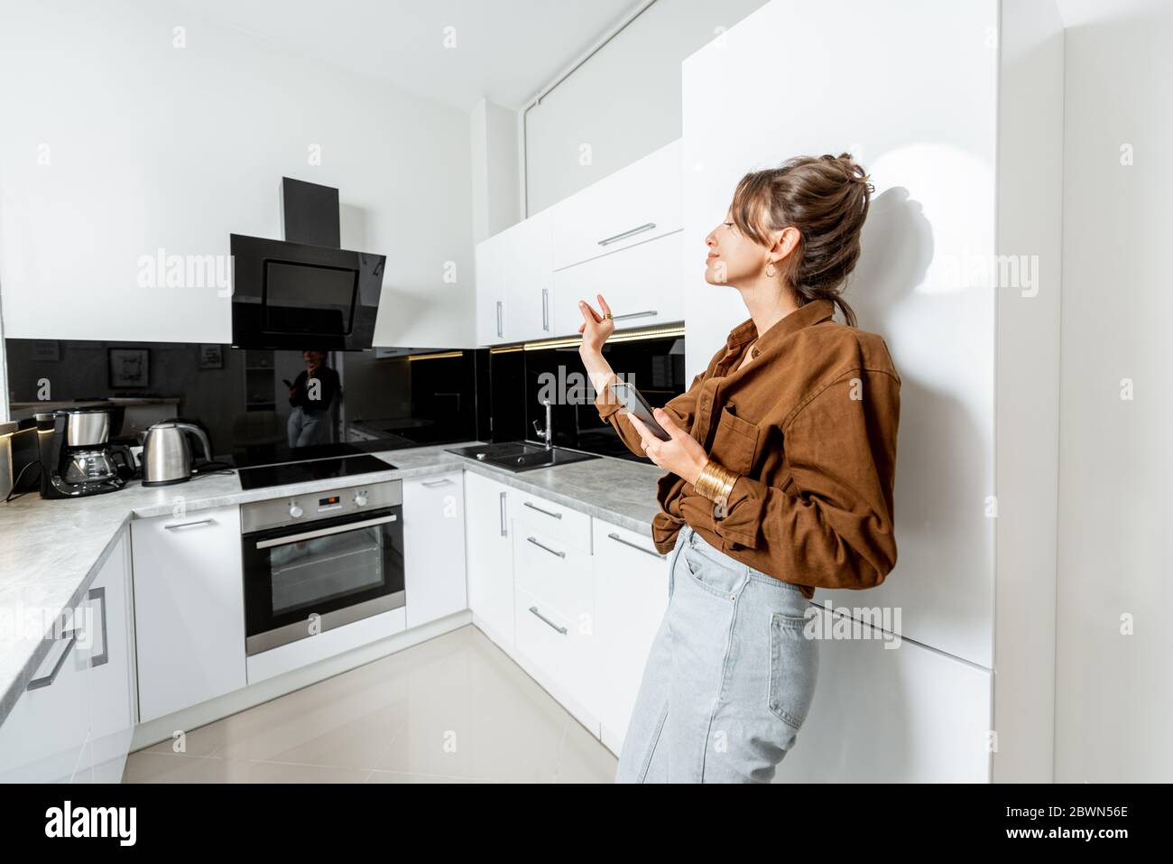 Young woman controlling kitchen appliances with mobile phone and voice commands, wide interior view. Smart home concept Stock Photo