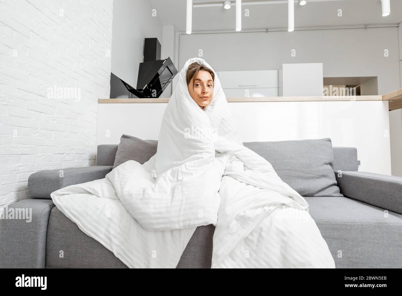 Sleepy woman covered with a white blanket on the couch at home, wide interior view. Concept of self-isolation and flu diseases at home Stock Photo