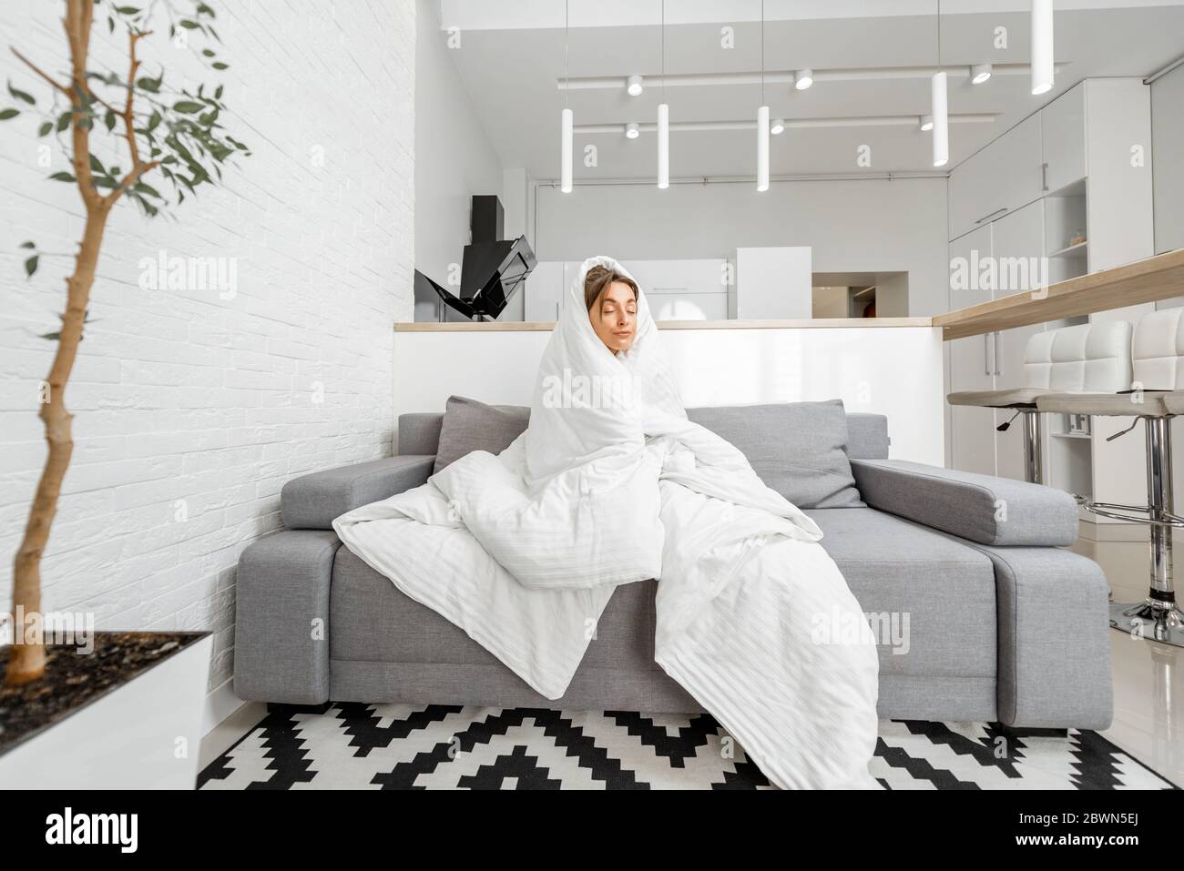 Sleepy woman covered with a white blanket on the couch at home, wide interior view. Concept of self-isolation and flu diseases at home Stock Photo