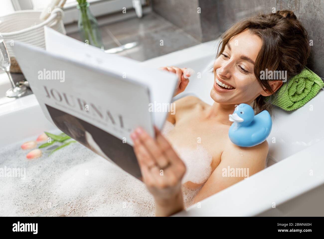 Young and cheerful woman relaxing while taking a bath, lying with rubber duck and reading some magazine at home Stock Photo