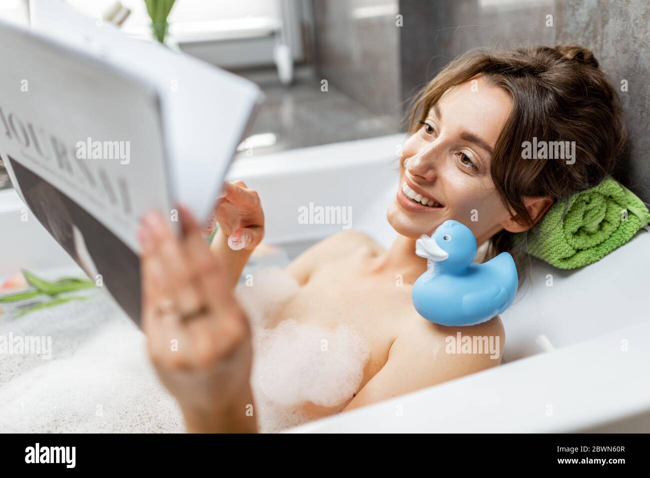 Young and cheerful woman relaxing while taking a bath, lying with rubber duck and reading some magazine at home Stock Photo