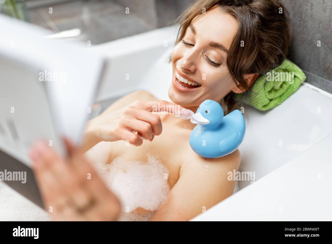 Young and cheerful woman relaxing while taking a bath, lying with rubber duck and reading some magazine at home Stock Photo