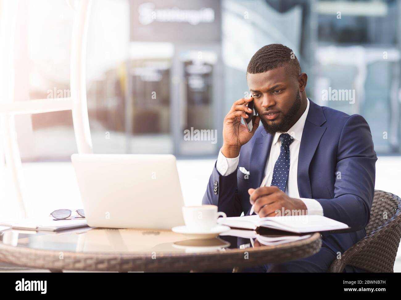 African Business Guy Multitasking Talking On Cellphone Working On Laptop And Taking Notes Sitting In Outdoor Cafe. Busy Lifestyle Concept Stock Photo