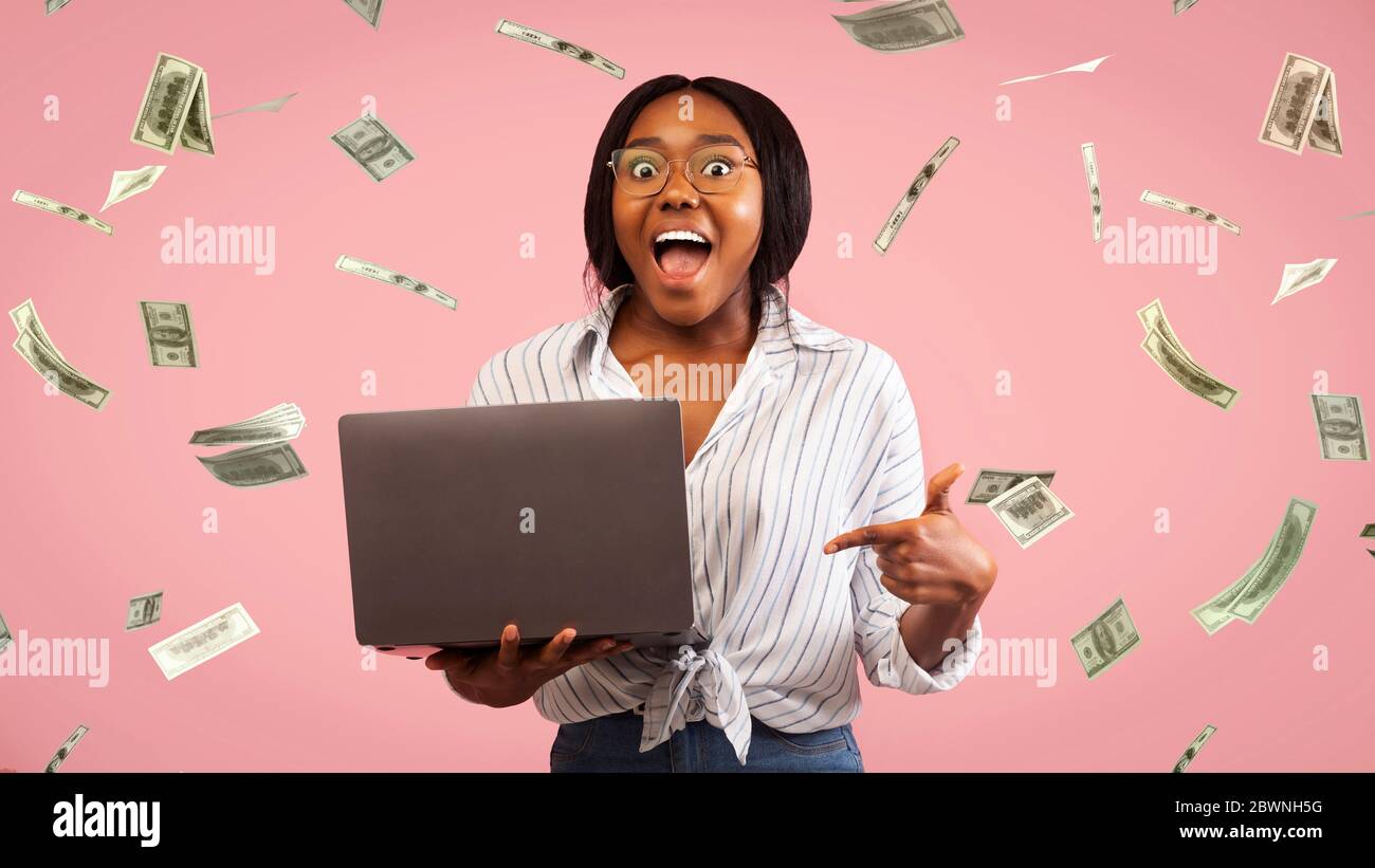 Online Casino Winner. Creative Collage Of Euphoric Black Girl Standing With Laptop Under Money Shower Of Falling Dollar Banknotes, Panorama Stock Photo