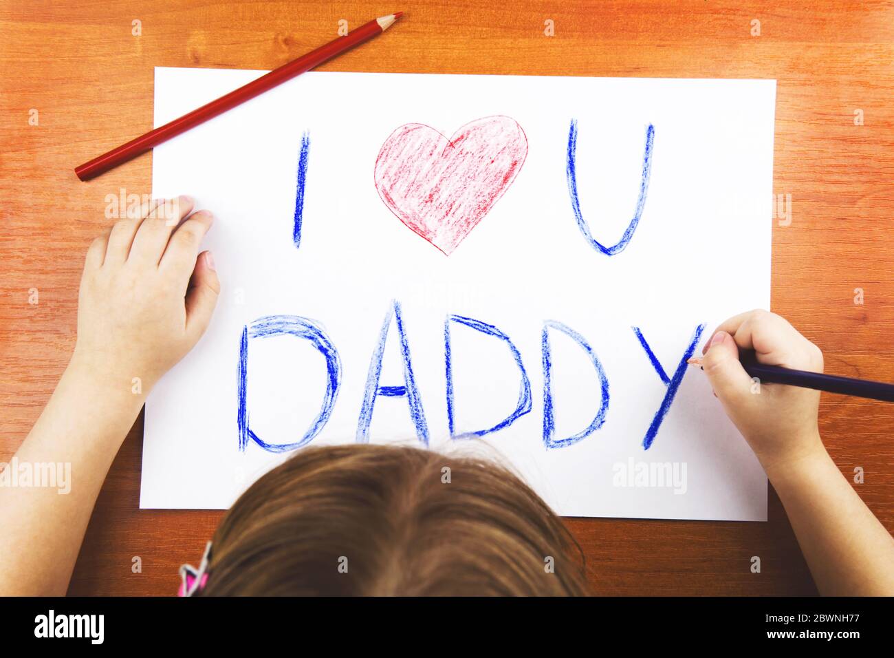 Fathers day concept. Girls hands drawing card with words I Love U Daddy, and red heart, and color pencils on wooden desk. Stock Photo