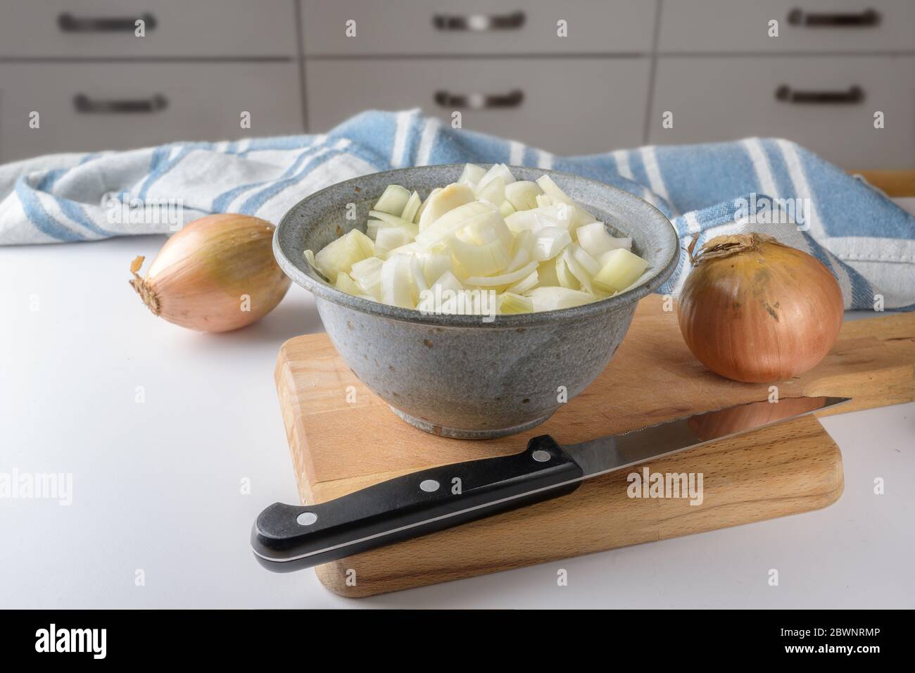 Onions whole and chopped in a bowl, knife and cutting board on a white table in the kitchen, selected focus Stock Photo