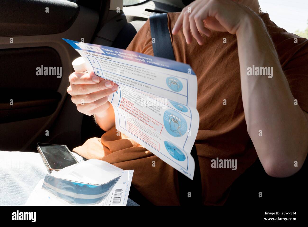 A young man in his twenties reads printed instructions after inserting a swab into his mouth to reach his tonsils and into his nasal passage from the rear seat of a car for a self-administered Coronavirus (COVID-19) test in south London. There are four steps to the self-administered Covid-19 test (inserting a swab into the nose and throat) which the public works through in their car, windows up and all communications with army personnel via phone, in a south London leisure centre, on 2nd June 2020, in London, England. The kit provided consists of a booklet, plastic bag, swab, vial, bar codes a Stock Photo