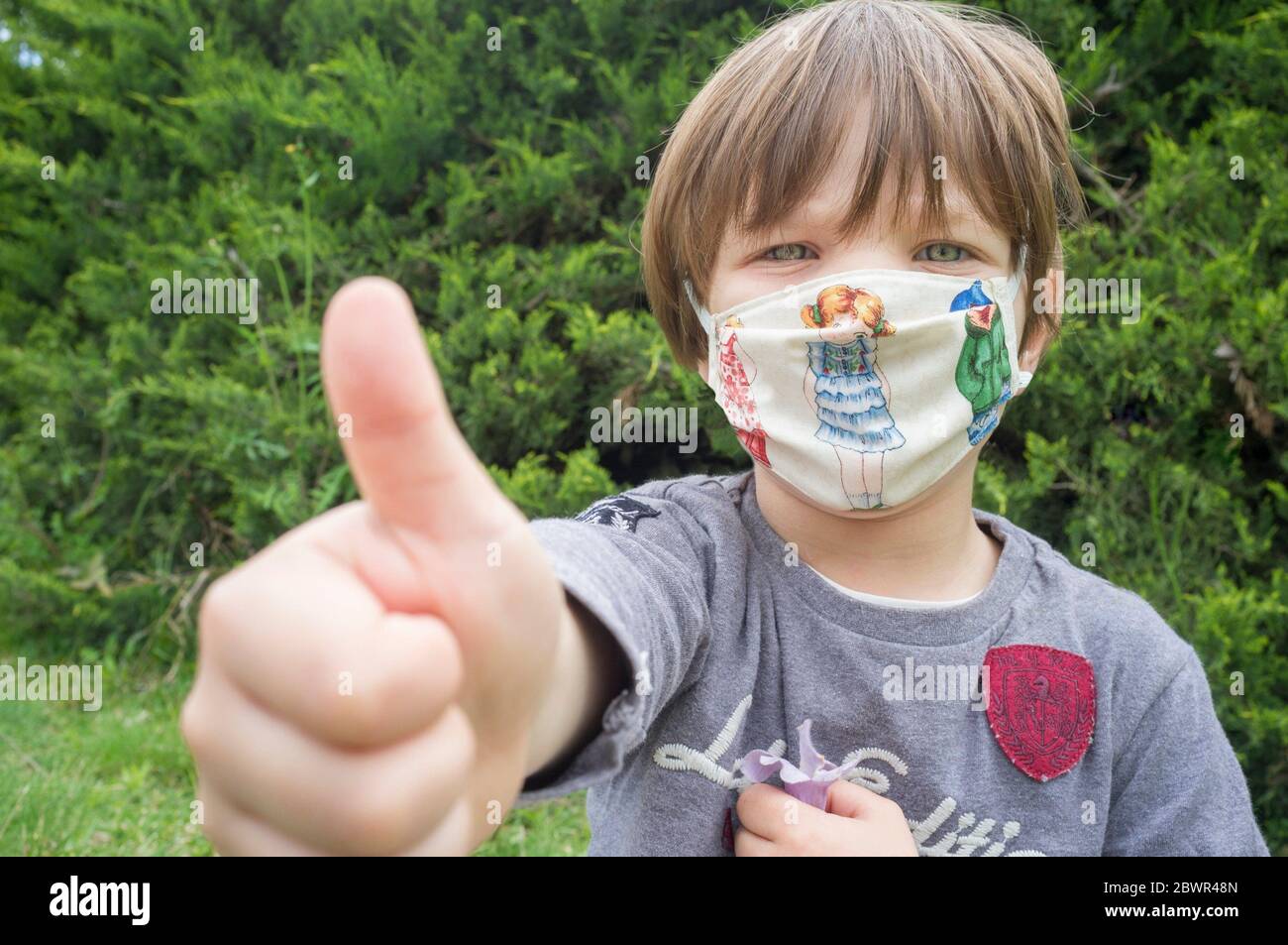 4 years little boy wearing a face mask print with children motifs. He is dooing the thumbs up gesture. Stock Photo