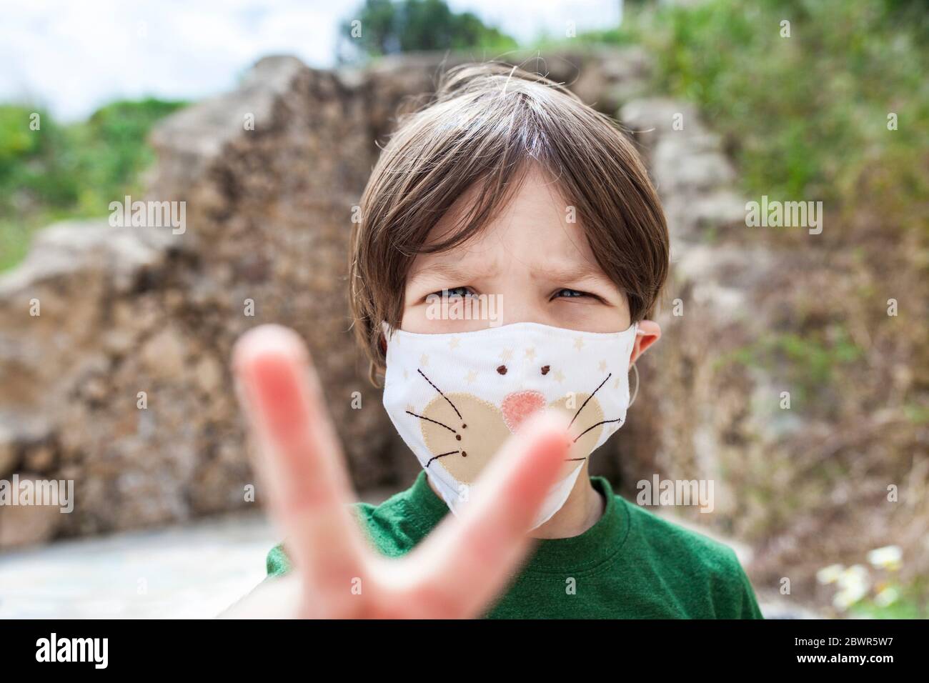 Little boy holds up two fingers for victory over Covid-19 Pandemic. He is wearing face mask with children motifs. Stock Photo