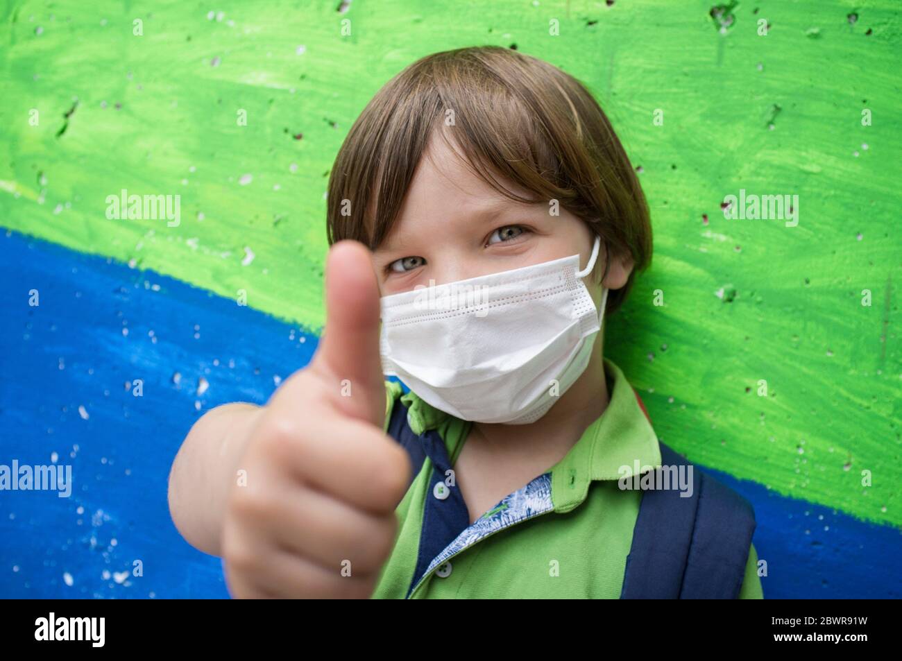 4 years little boy wearing a face mask doing the thumbs up gesture. Lively colored concrete block as background. Stock Photo
