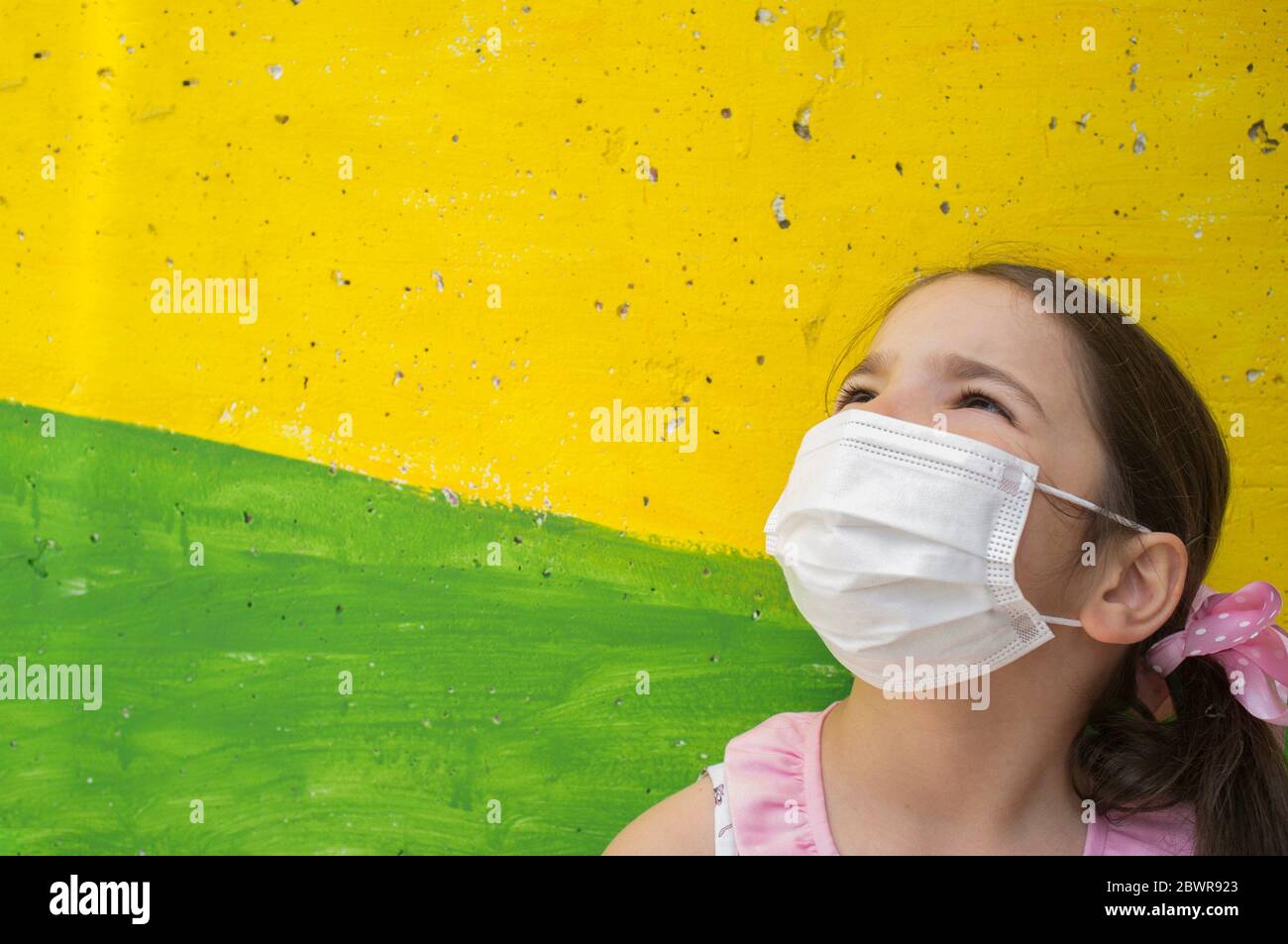 Hopeful little girl wears face mask during Covid-19 pandemic. Lively colored concrete block as background. Stock Photo