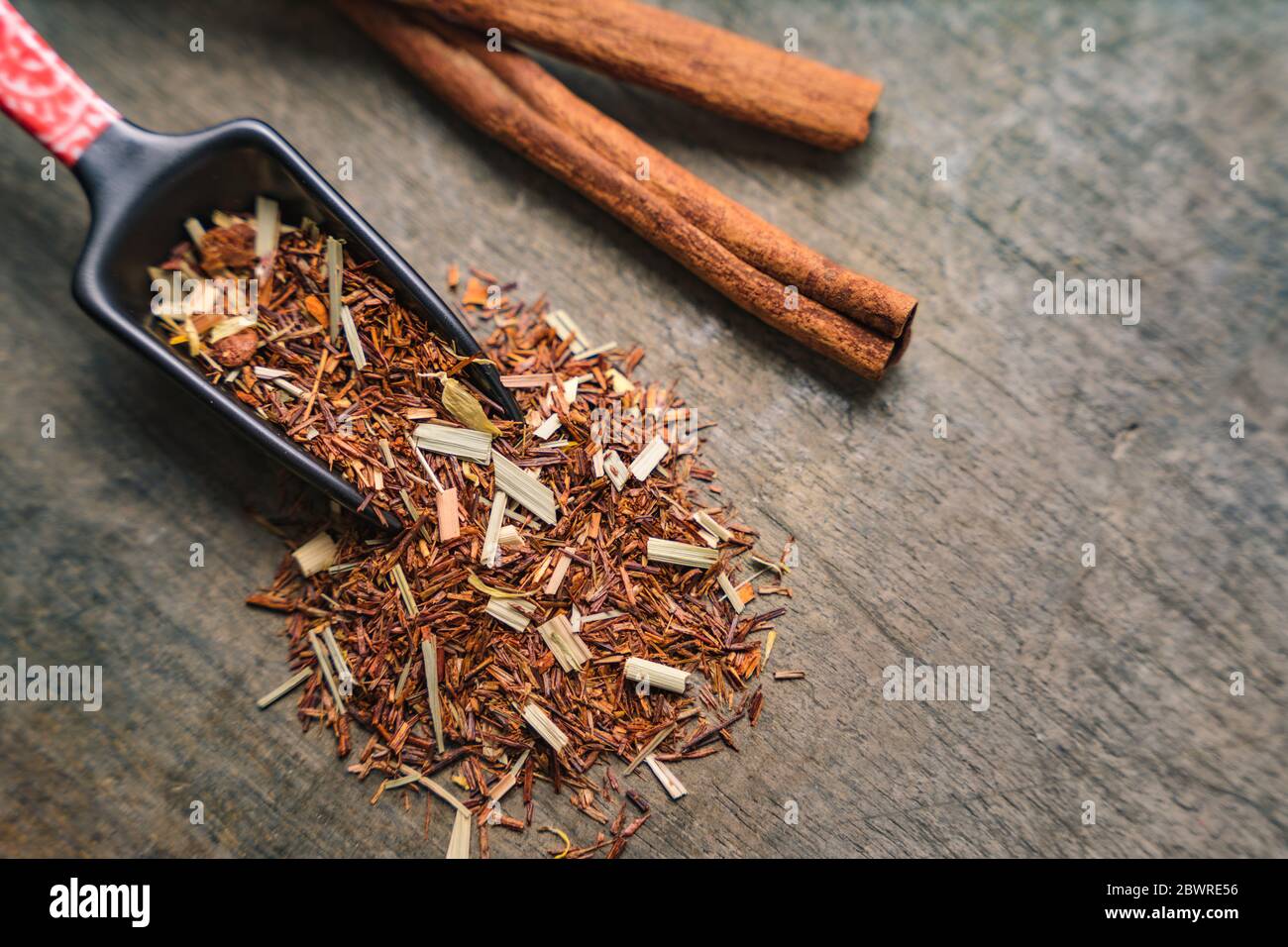 Japanese measuring tea spoon spreads aromatic rooibos dried leaves on wooden surface. Cinnamon bark strips on the side Stock Photo