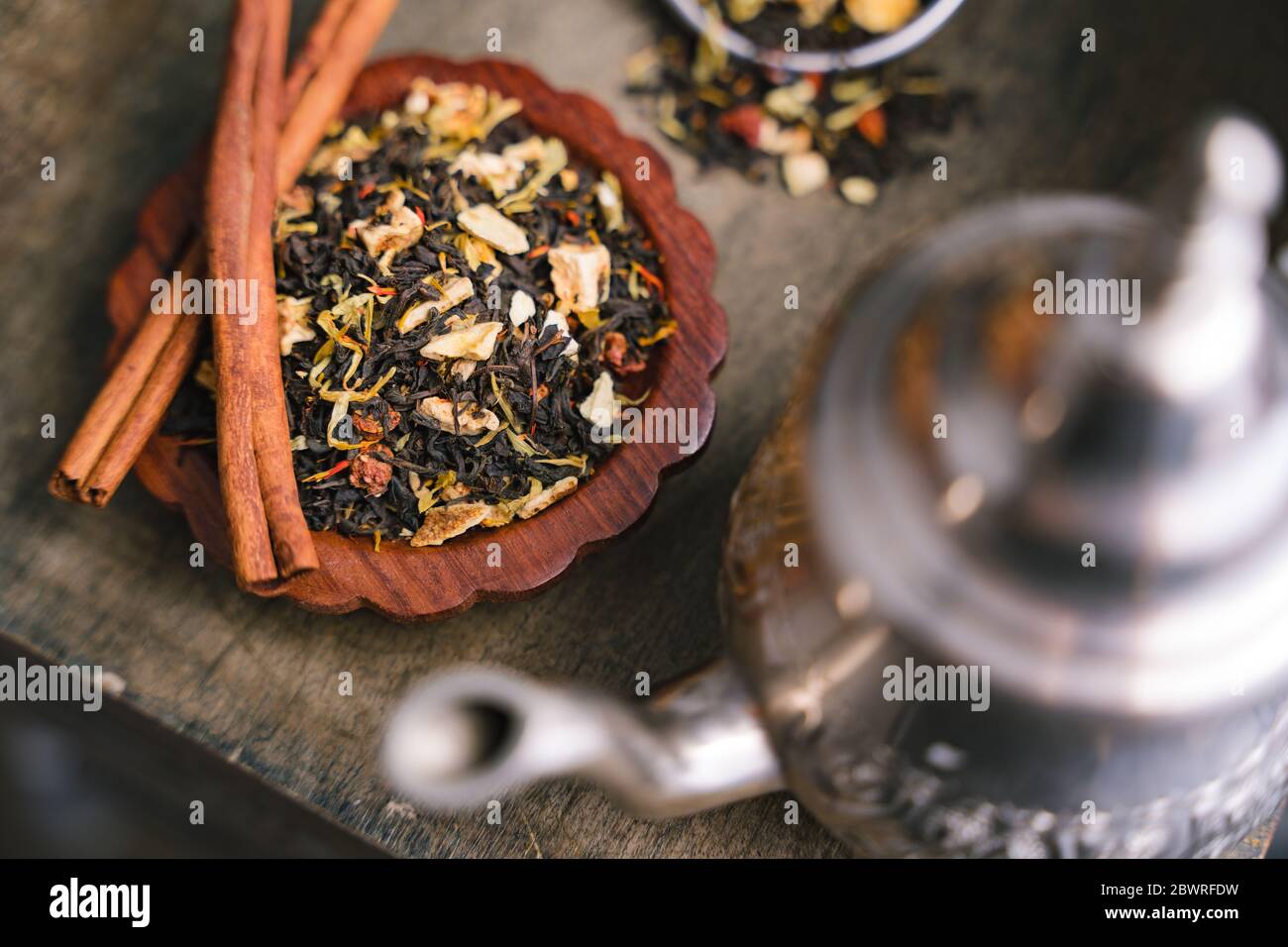 Aromatic fruity black tea dried leaves and cinnamon bark strips laid on small wooden tray with metal kettle in foreground Stock Photo