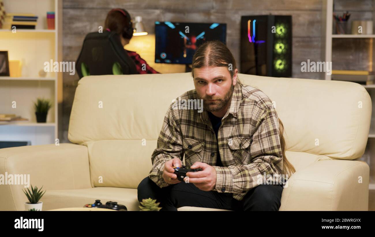 Focused bearded man sitting on couch playing video games using wireless controller. Girlfriend playing on computer in the background. Stock Photo