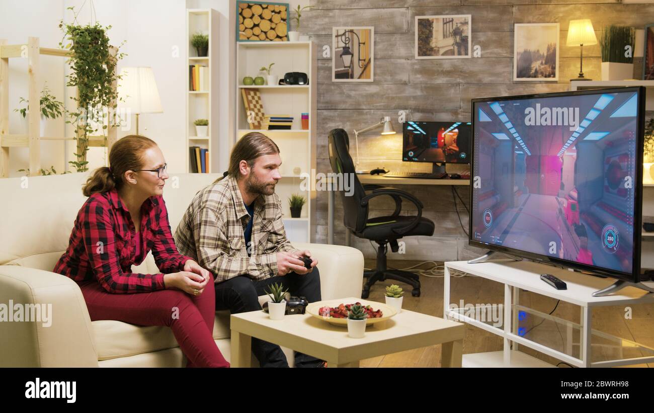 Cheerful girlfriend talking with her boyfriend while he's playing video games on tv using wireless joystick. Stock Photo