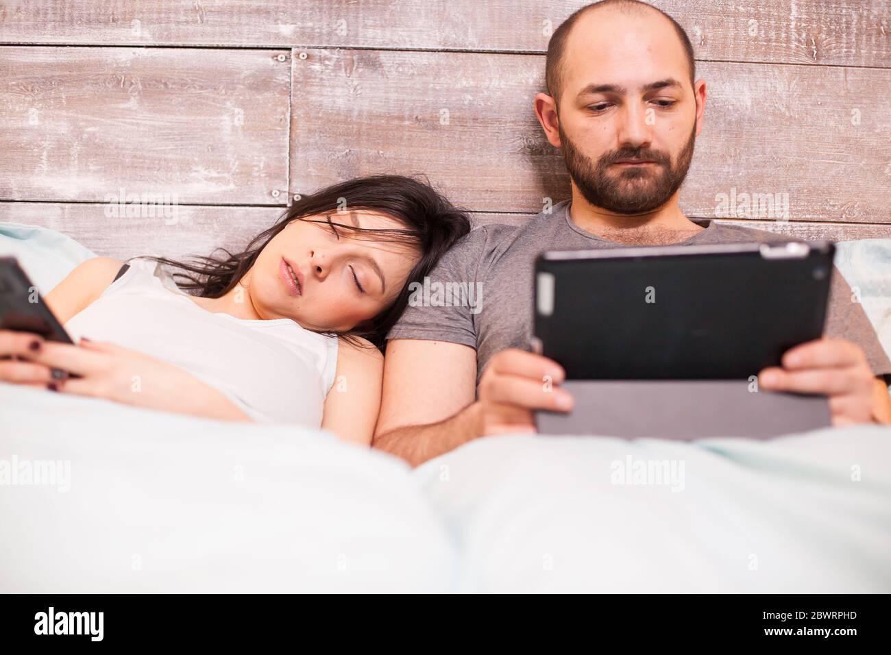 Beautiful wife in pajamas sleeping while her husband is working on tablet computer. Stock Photo