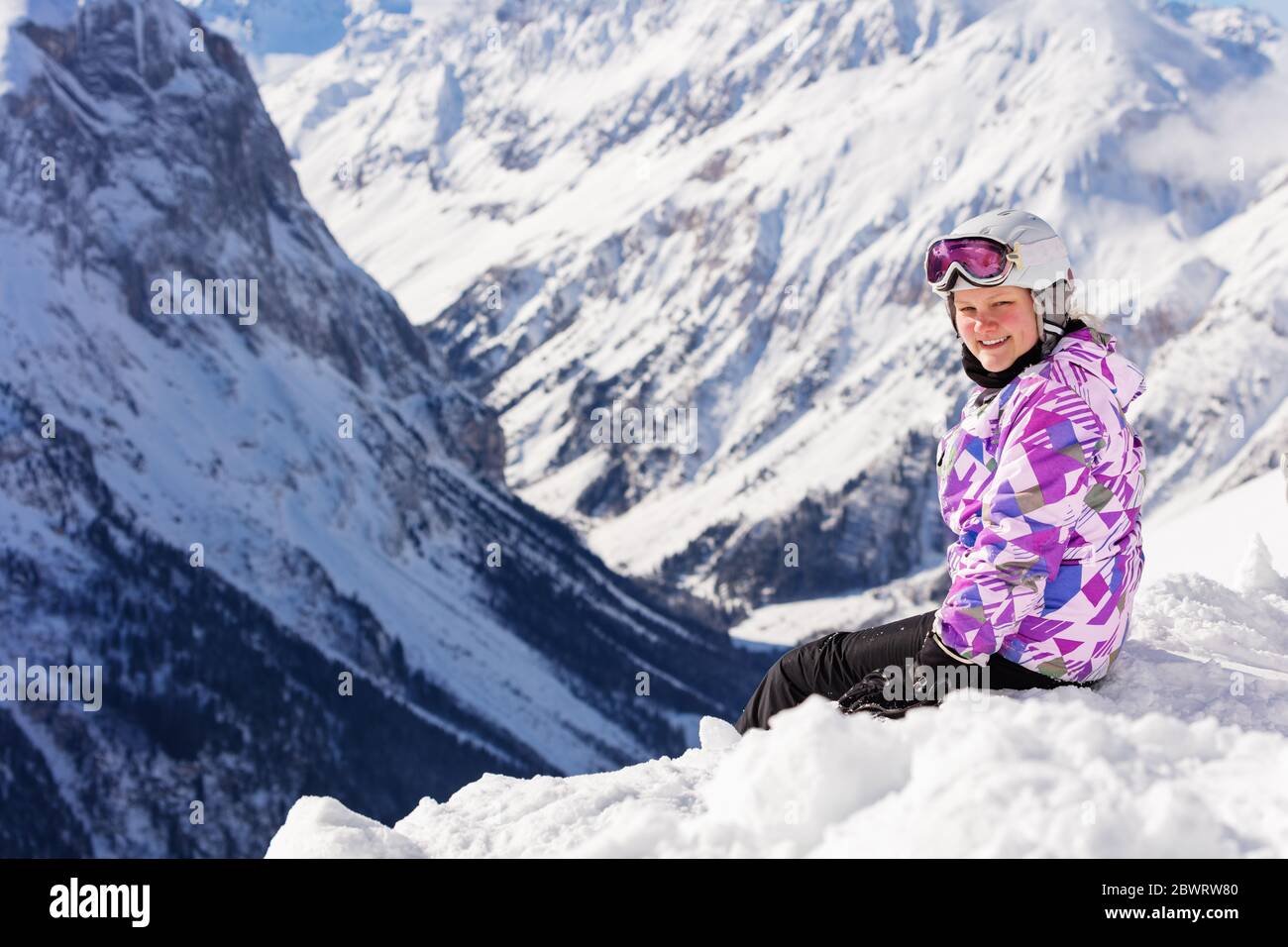 Cute teen girl sit in snow on top of the mountain in ski outfit with helmet and mask turn back smiling to camera Stock Photo