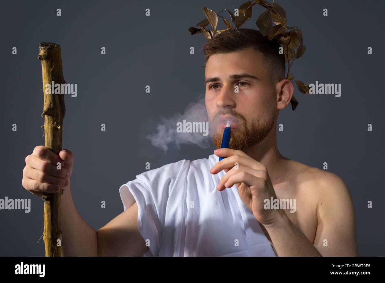 a bearded god smokes an electronic cigarette. A young guy in the guise of an ancient Greek god smokes a modern cigarette. Dramatic portrait on a gray Stock Photo