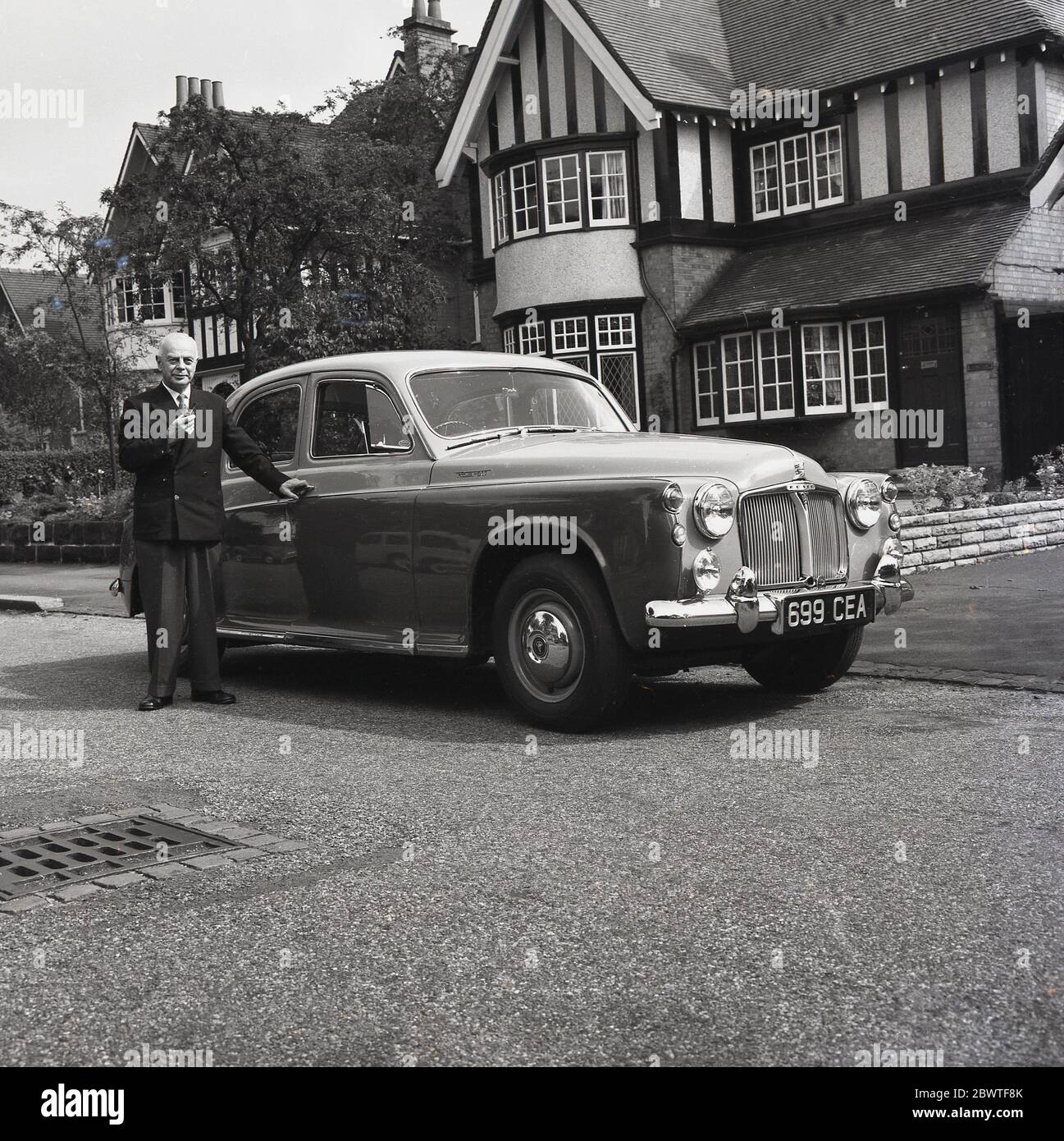 1961, historical, an elderly English gentleman in a balzer and slacks and holding a pipe, standing proudly by his two-tone Rover 100 motorcar outside a large house in the suburbs, England, UK. Designed by Gordon Bashford and from of the Rover P4 family of cars - known as 'Auntie Rovers', -these elegant motorcars were driven by Royalty- Grace Kelly owned one - and are an important part of British culture. The Rover 100 was introduced in October 1959 and was a 4-door saloon with a luxurious with a leather and wood interior. Stock Photo