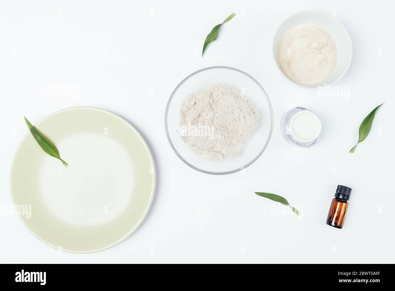 Ingredients for homemade beauty treatments, natural clay mask, moisturizer cream and aroma essence next to green leaves on white table, top view. Stock Photo