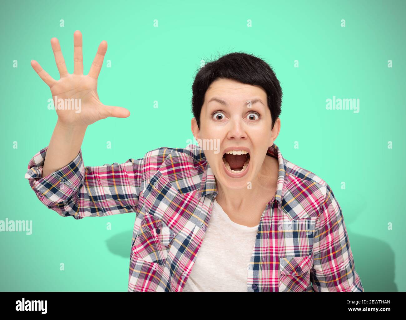 Angry woman isolated on neo mint background. Beautiful furious short haired brunette dressed casual plaid shirt shouting and waving her palm hand Stock Photo