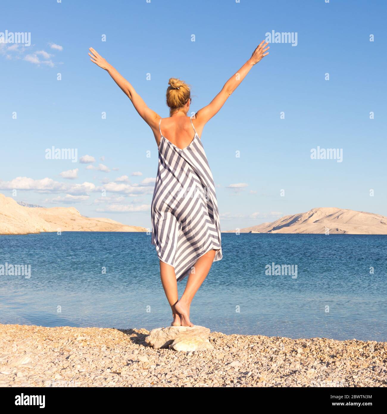 Happy carefree woman rising arms, wearing beautiful striped summer dress enjoying late afternoon on white pabbled beach on Pag island, Croatia Stock Photo