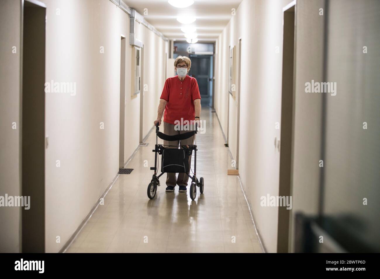 Senior woman wearing mask and walking with wheeled walker in corridor ...