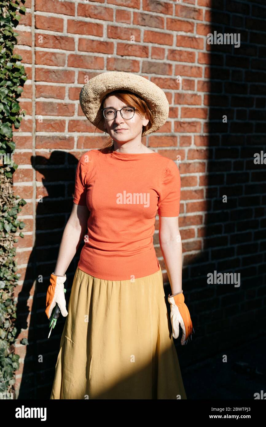 Portrait of mature woman with gardening gloves and pruner standing in front of brick wall Stock Photo