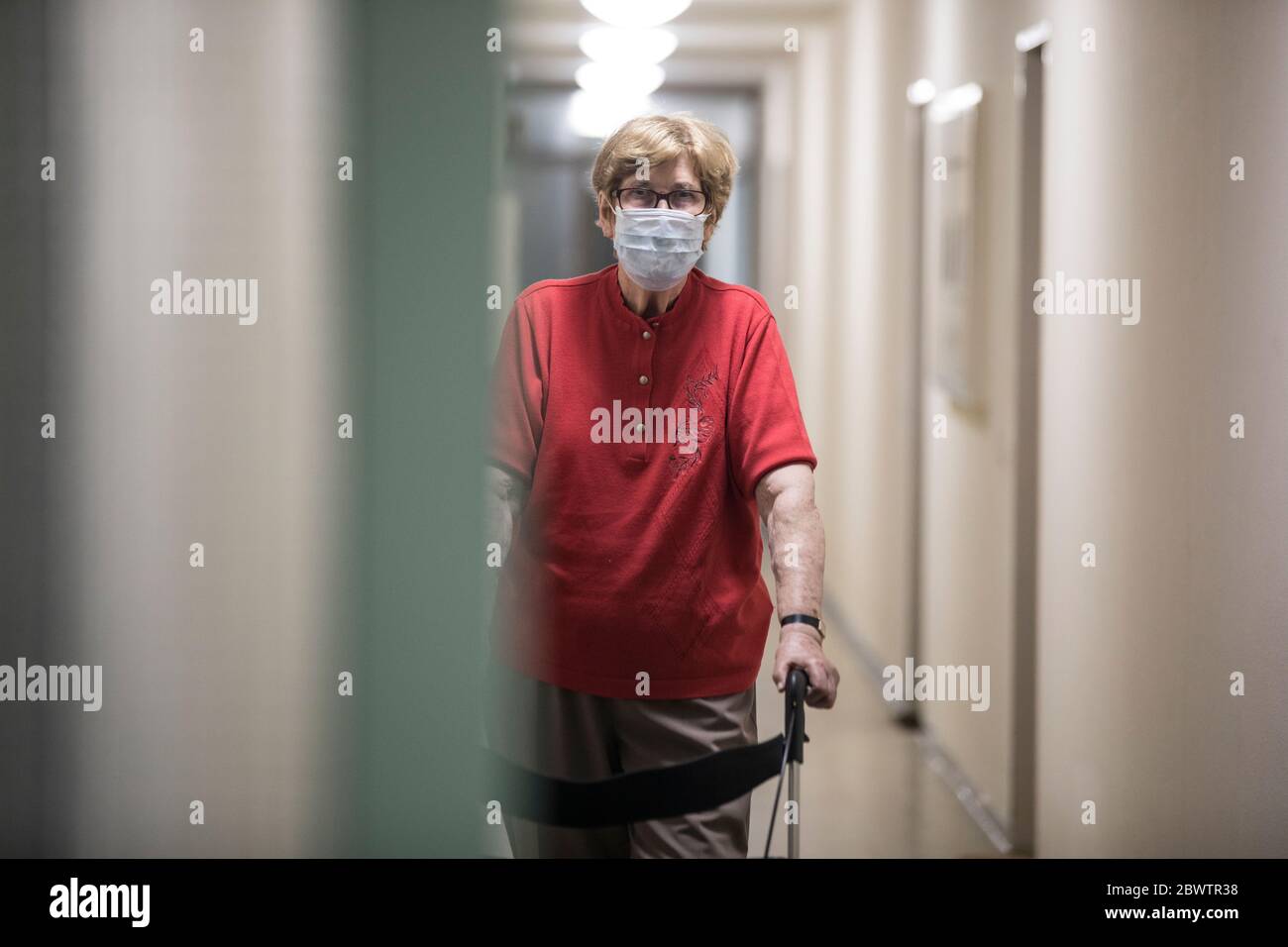 Senior woman wearing mask and walking with wheeled walker in corridor ...