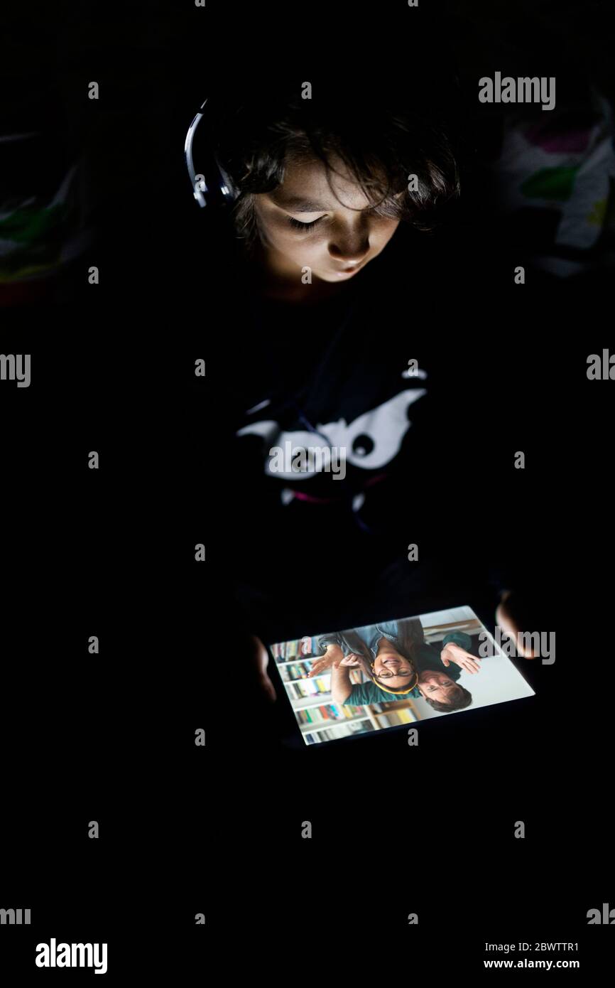 Boy sitting in his dark room at home using headphones and digital tablet for video chat Stock Photo
