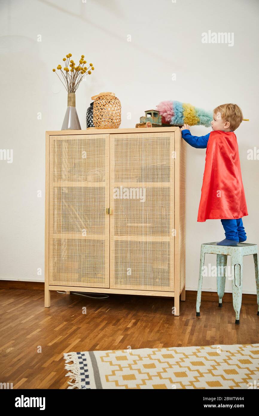 Boy wearing superman costume and cleaning toy with feather duster on a sideboard at home Stock Photo