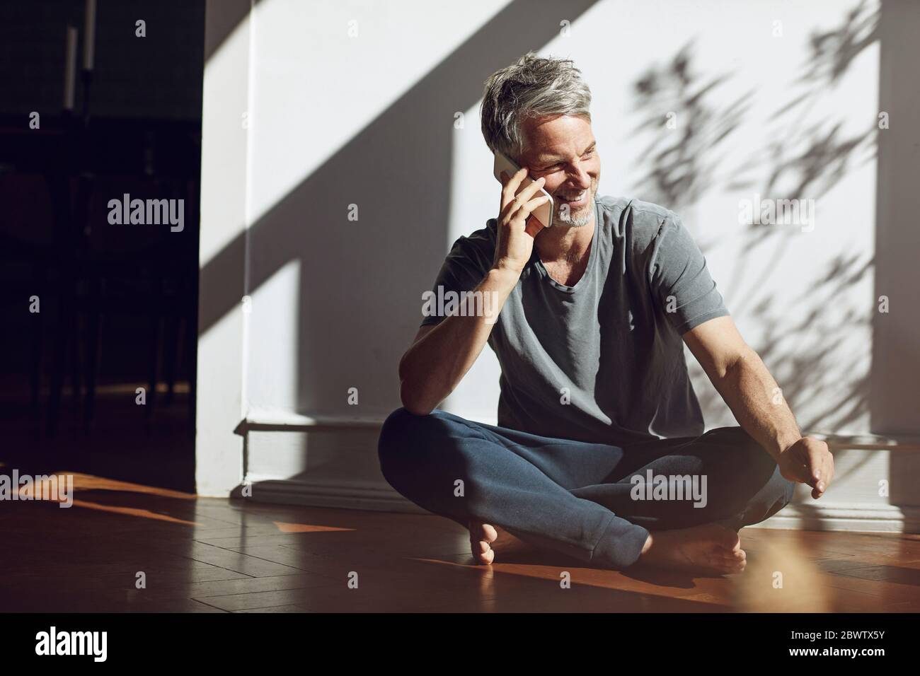 Smiling mature man sitting on the floor at home talking on the phone ...