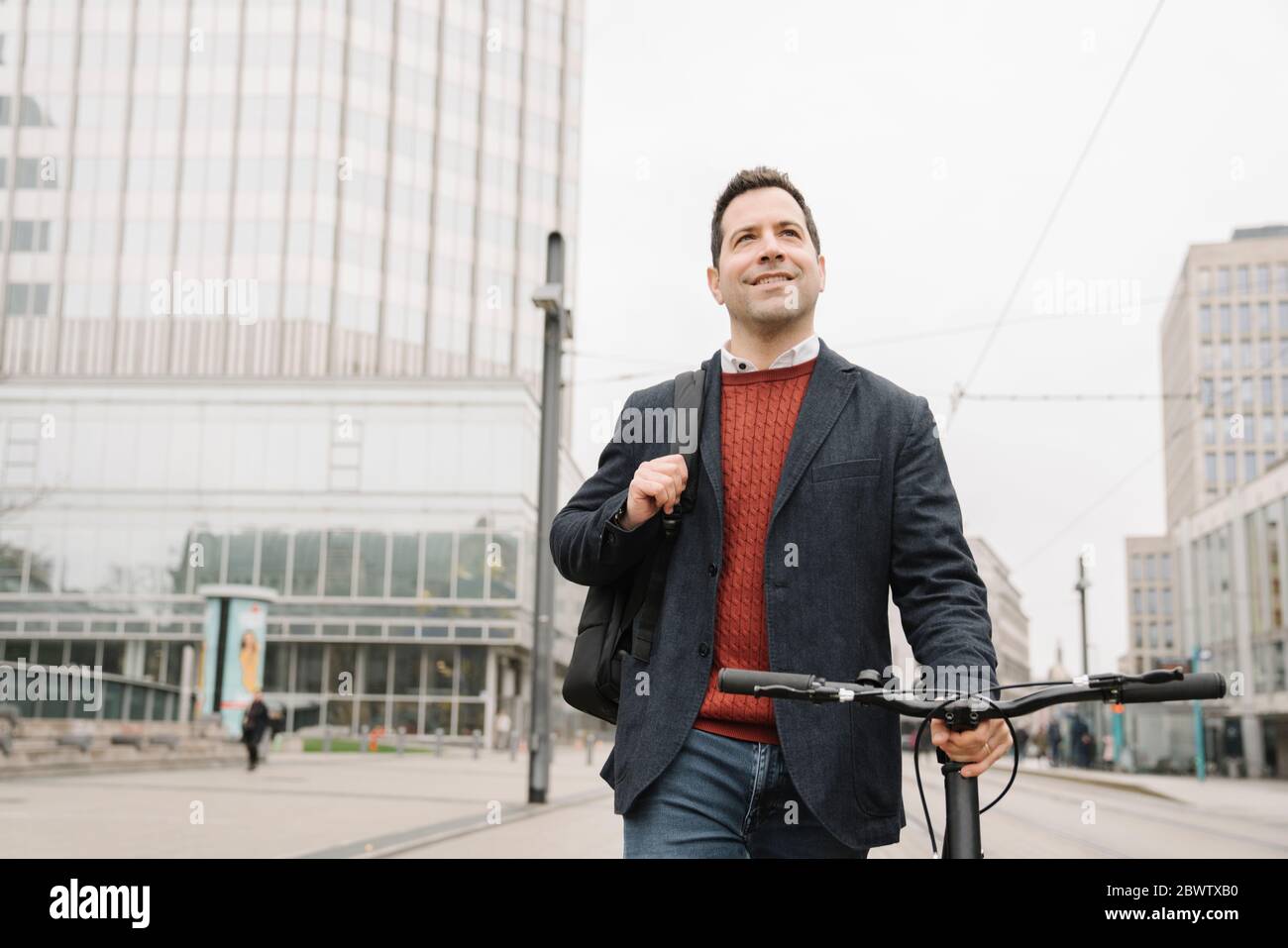 Smiling businessman looking away while walking with bicycle on city street in Frankfurt, Germany Stock Photo