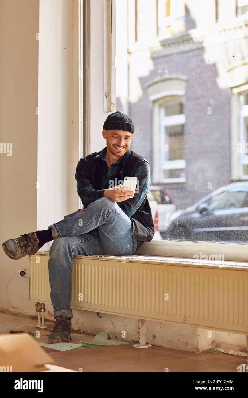 Man refurbishing shop location, sitting on windowsill, using smartphone Stock Photo