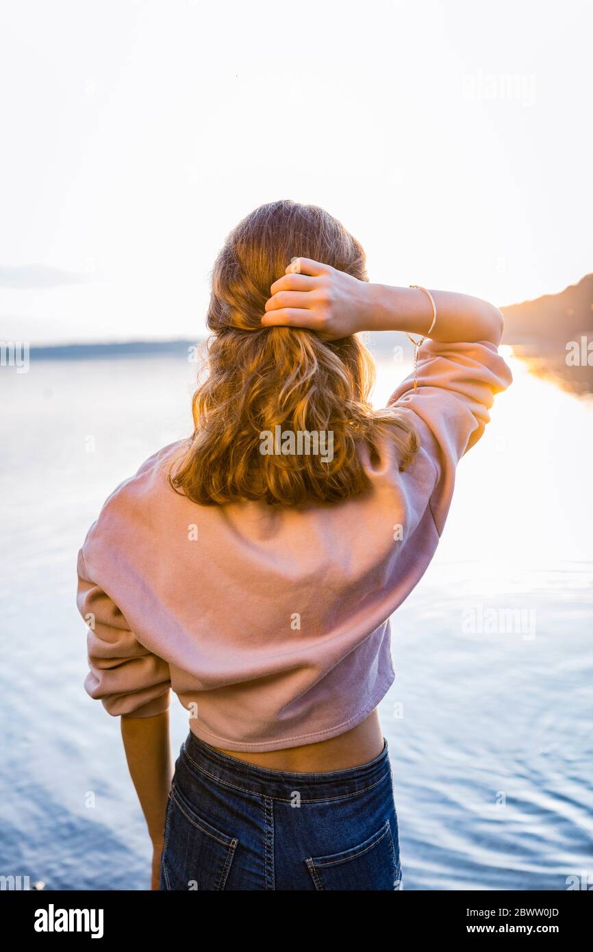 Rear view of girl with hand in hair looking at lake during sunset Stock Photo