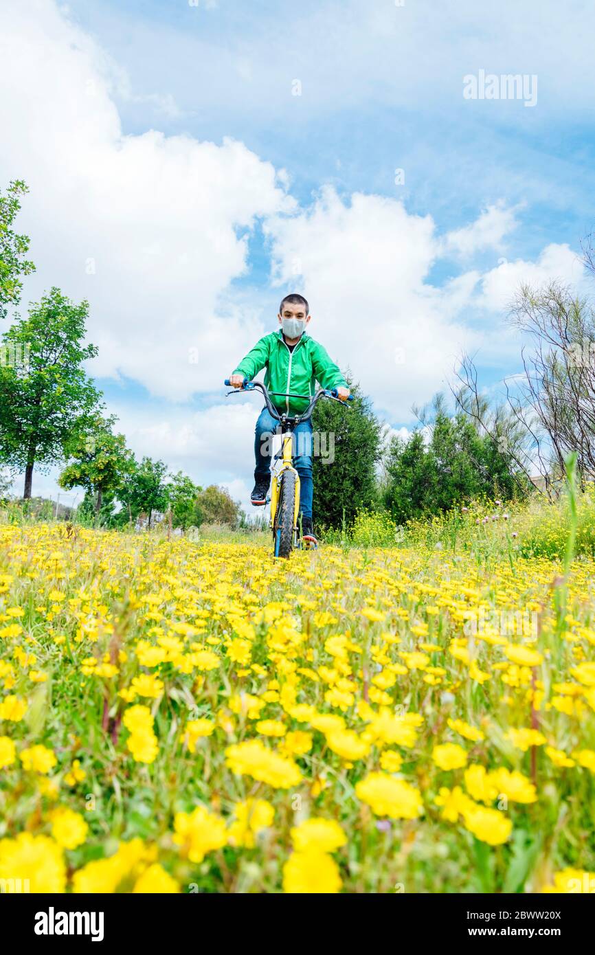 Full length of boy wearing face mask cycling over yellow flowers on field against sky Stock Photo