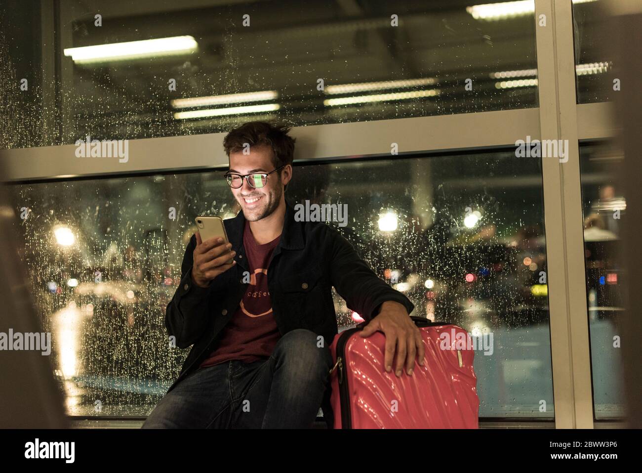Smiling young man with suitcase sitting at rainy window using smartphone Stock Photo