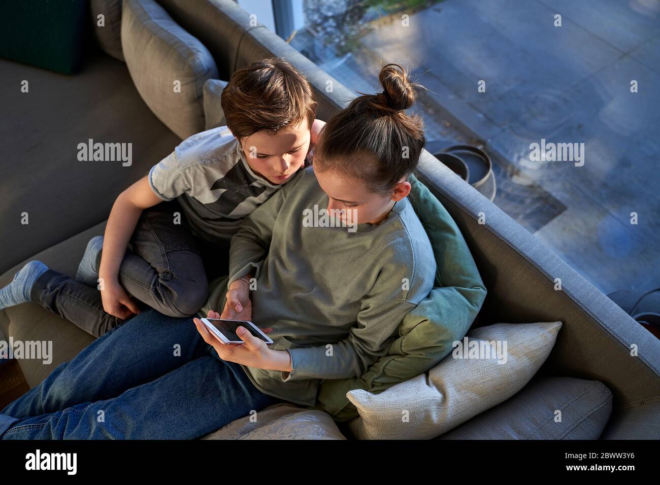Brother and sister sitting on couch at home using smartphone Stock Photo