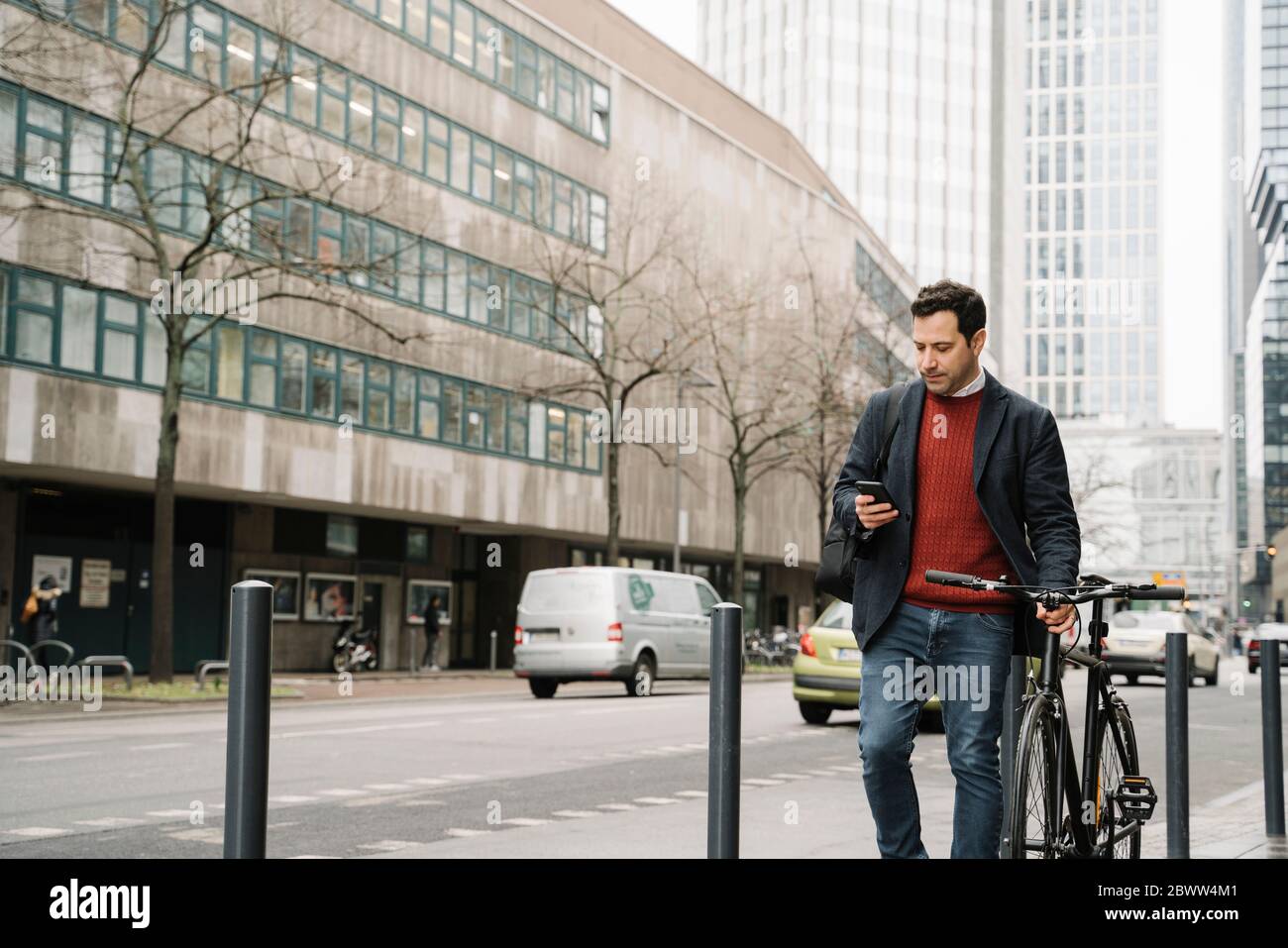 Businessman with bicycle using smart phone while walking against buildings in city, Frankfurt, Germany Stock Photo