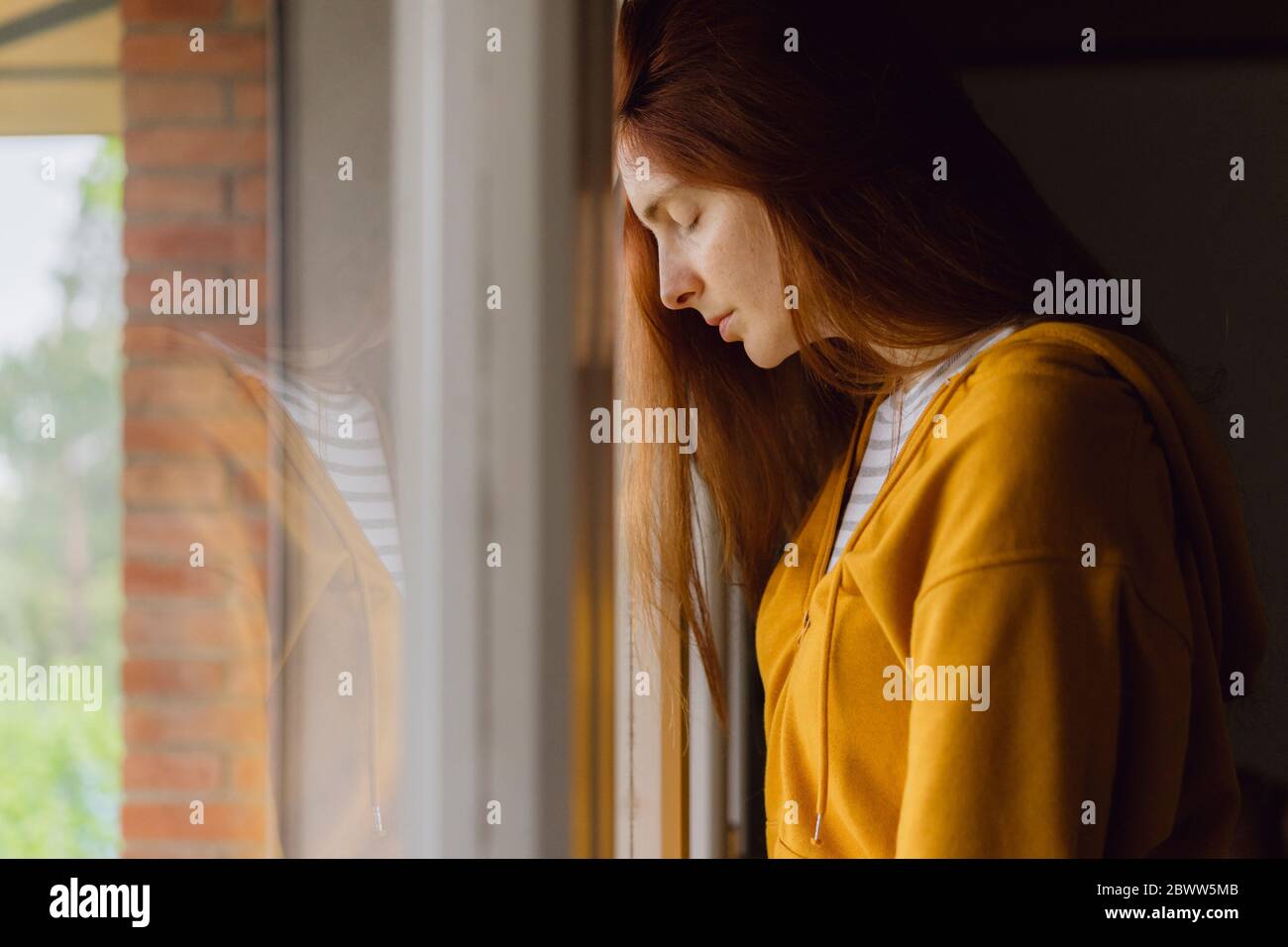 Redheaded woman with eyes closed leaning against window Stock Photo