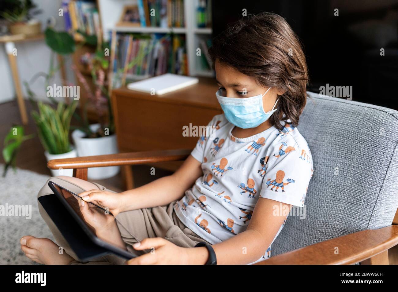 Boy with surgical mask sitting on armchair at home using digital tablet Stock Photo